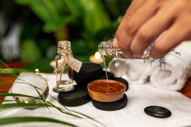 A man pours oil from a jar of essential oils standing on stones for a therapy stone and located on a terry towel next to which are transparent spheres, a twisted terry towel of white color and a sprig of lavender